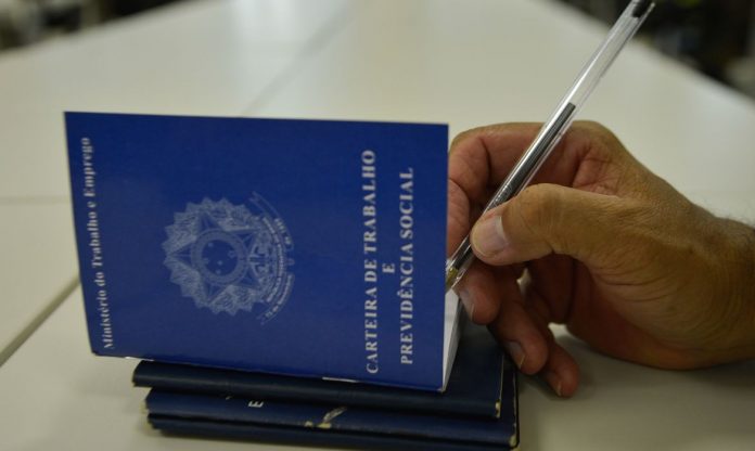 #Pracegover Foto: Na imagem há algumas carteiras de trabalho e uma pessoa com uma caneta na mão assinando o documento