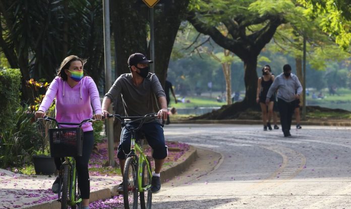 #Pracegover Foto: na imagem há uma rua asfaltada, árvores, pessoas caminhando e duas pessoas andando de bicicleta