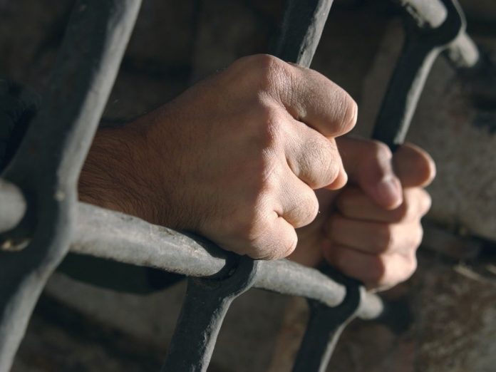 #Pracegover Foto: na imagem há um preso com as mãos em uma grade
