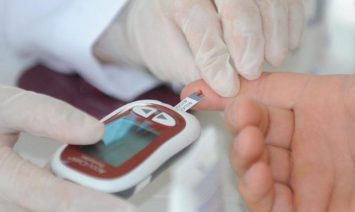#Pracegover Foto: Uma pessoa com luvas e um aparelho realiza um teste de diabetes em uma paciente