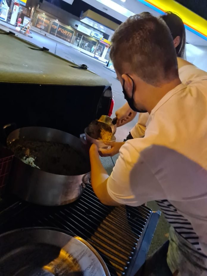 #Pracegover Foto: na imagem há um homem preparando o alimento e colocando em um recipiente. Há também uma mulher que segura esse recipiente e um carro