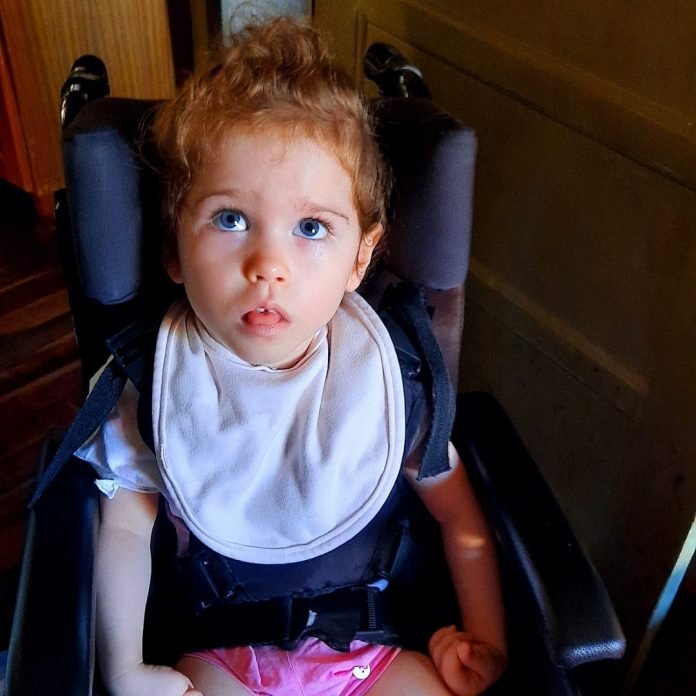 Helena #Pracegover Foto: na imagem há uma criança, uma menina de 2 anos, com babador, blusinha azul e short rosa