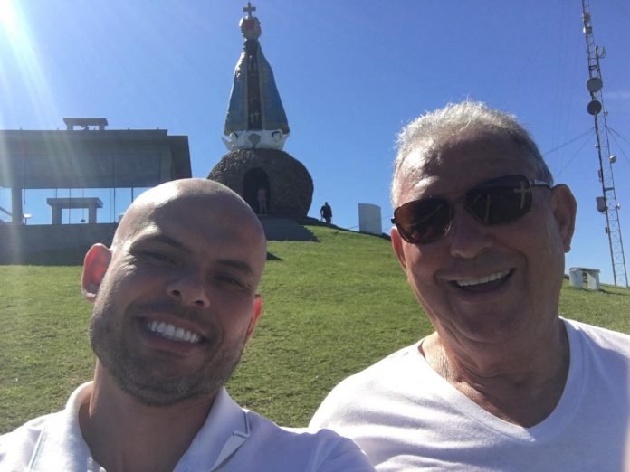 #Pracegover Foto: na imagem há dois homens sorridentes e no fundo há também há imagem de Nossa Senhora Aparecida