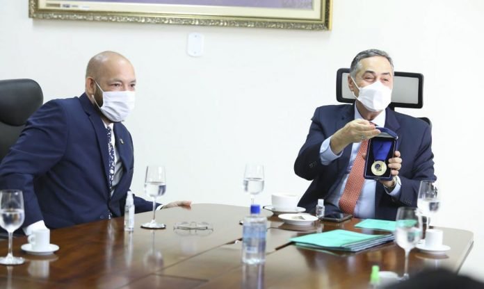 tse_ministro_luis_roberto_barroso #Pracegover Foto: na imagem há dois homens, copos e garrafas de água, papéis, uma mesa e um dos homens segurando uma medalha