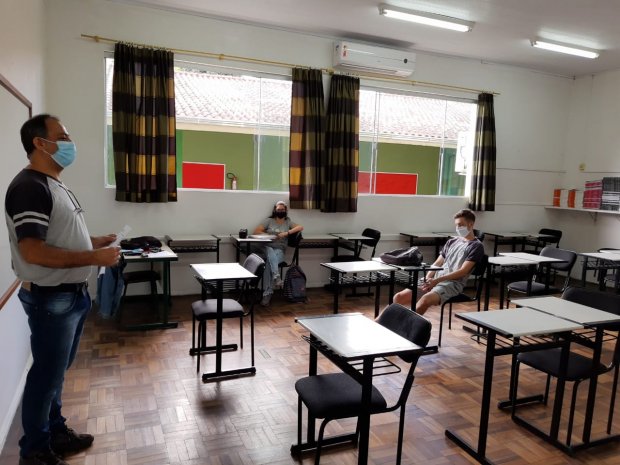 #Pracegover Foto: na imagem há uma sala de aula com várias carteiras, cortinas, janelas, um professor e dois alunos