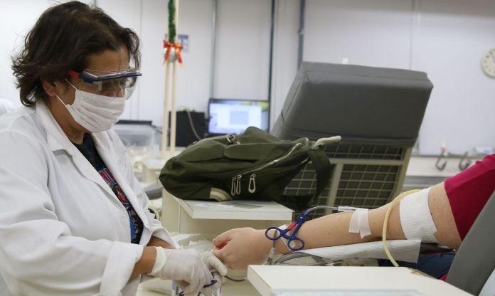 doação de sangue #Pracegover Foto: na imagem há uma mulher realizando o procedimento de captação em outra pessoa. Ela está de luvas e máscara. Em um balcão há uma mochila