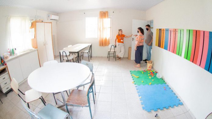 #Pracegover Foto: na imagem há três pessoas, cadeiras, mesa, tapetes, armário e brinquedos