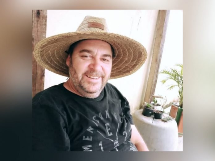 empresário #Pracegover Foto: na imagem há um homem sorrindente, de camiseta preta, chapéu. Próximo a ele há um celular e alguns plantas
