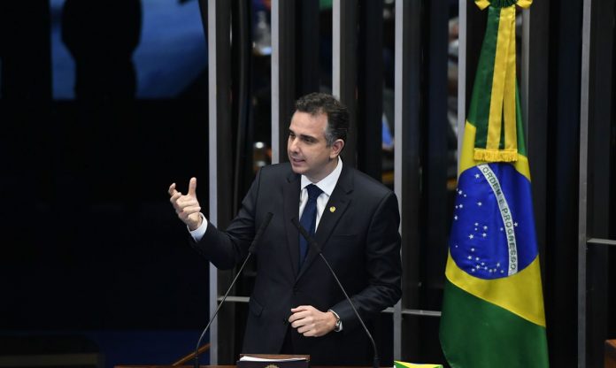 #Pracegover Foto: na imagem há um homem de terno, gesticulando com as mãos, microfones e a bandeira do Brasil