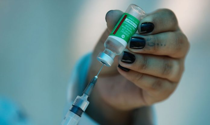 #Pracegover Foto: na imagem há uma mão com unhas pintadas, um frasco de vacina, uma seringa e agulha