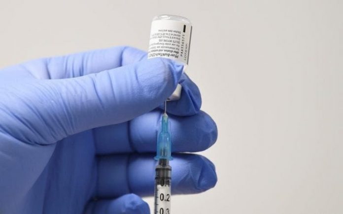 #Pracegover Foto: na imagem há um frasco de vacina, uma seringa com agulha e uma mão com luva azul