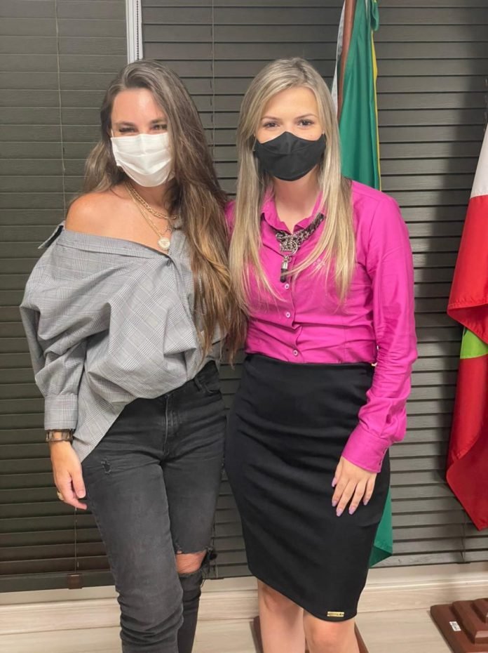 Bia e Paulinha #Pracegover Foto: na imagem há duas mulheres de máscara e hpróximo a elas há duas bandeiras, a do Brasil e de Santa Catarina