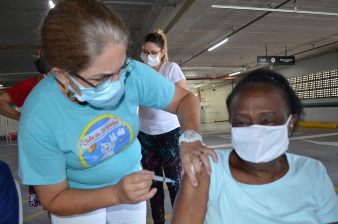 vacina tb #Pracegover Foto: na imagem há uma mulher aplicando a vacina em uma idosa e há outras duas pessoas aguardando