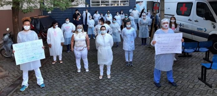 #pracegover Na foto, profissionais de saúde estão vestidos de branco, usando máscara, distantes um dos outros, com a mãos para trás, outros seguram cartazes