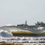 #pracegover Na foto, praia do Mar Grosso, em Laguna, com as ondas altas. Ao fundo o farol dos molhes