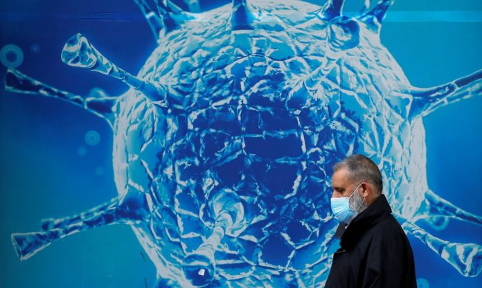 #Pracegover Na foto, homem em frente a um painel grande com uma imagem de uma célula do novo coronavírus