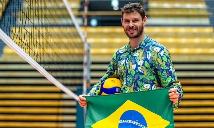 #Pracegover Foto: na imagem há um homem, uma bola, a bandeira do Brasil, uma rede de vôlei e arquibancada