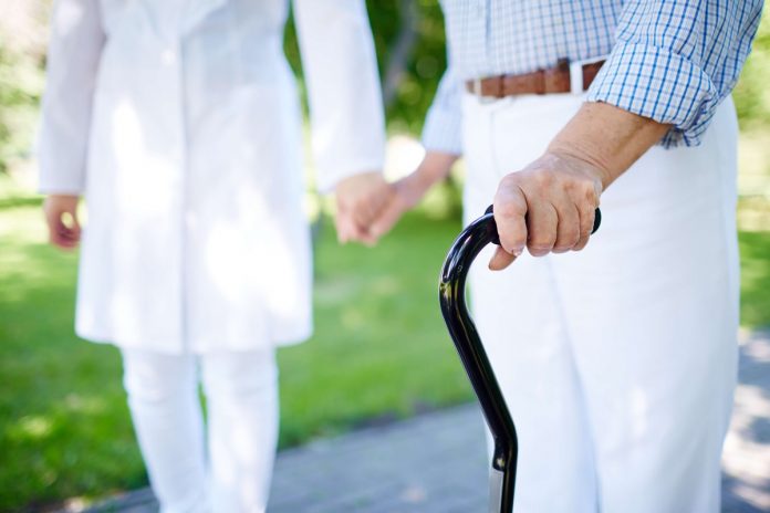 #Pracegover foto: na imagem há um idoso caminhando com o auxilio de uma profissional de saúde e uma bengala