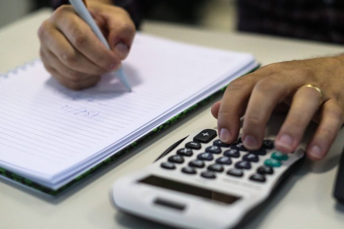 #Pracegover foto: na imagem há mãos, caneta, folhas e uma calculadora