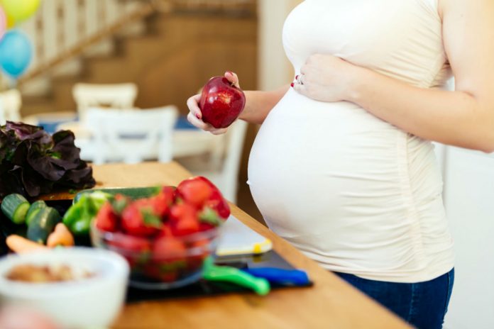 #Pracegover foto? na imagem há uma mulher grávida com a mão na barriga, na outra mão uma maça e frutas na mesa