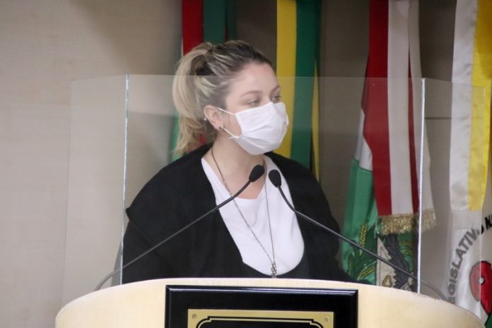 #Pracegover foto: na imagem há uma mulher com máscara, bandeiras, microfone e tribuna