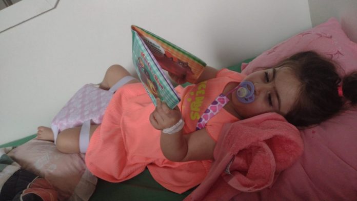 #Pracegover foto: na imagem há uma menina deitada, segurando um livro e com chupeta
