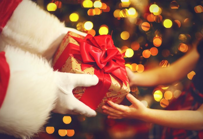#Pracegover foto: na imagem há mãos, presente e uma árvore de Natal