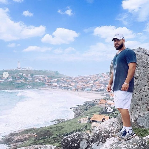 #Pracegover Foto: na imagem há um homem de bermuda nranca, camiseta azul e boné claro. Há o mar, montanhas, pedras e casas