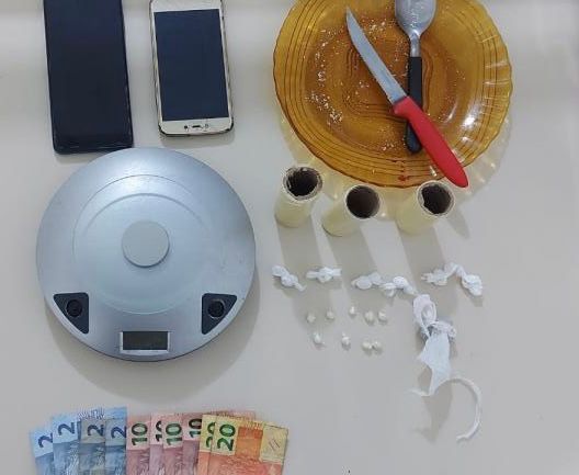 #PraCegoVer Na foto, há dois aparelhos de telefone celular, drogas, dinheiro uma balança de precisão e materiais utilizados para porcionar e embalar os entorpecentes