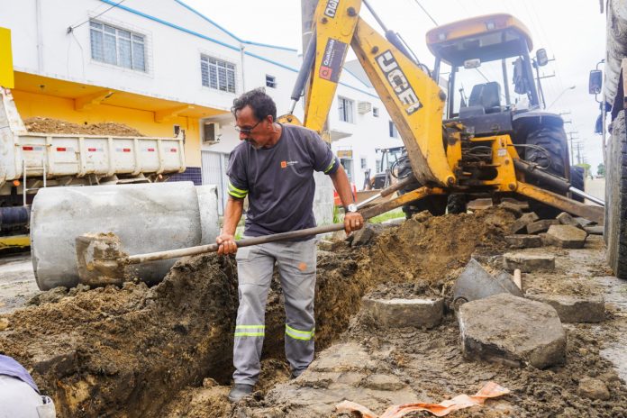 #PraCegoVer Na foto, um homem com uma pá cava um buraco na terra. Ao fundo há uma escavadeira