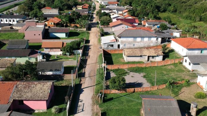 #PraCegoVer Na foto, a imagem área de uma estrada sem pavimentação circundada por várias casas e pequenos prédios