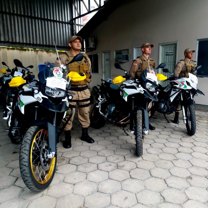 #PraCegoVer Na foto, três policiais militares estão parados ao lado de três motocicletas
