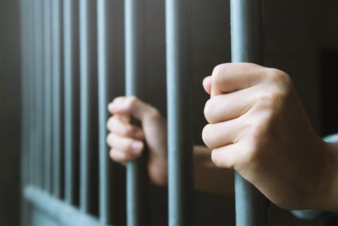 #PraCegoVer Na foto, uma pessoa com as mãos entre as grades de uma prisão
