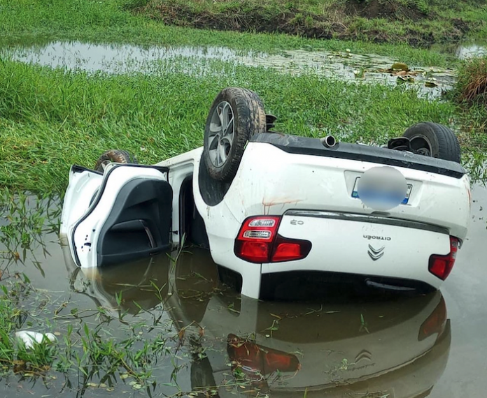 #PraCegoVer Na foto, um carro capotado e parcialmente submerso em uma vala