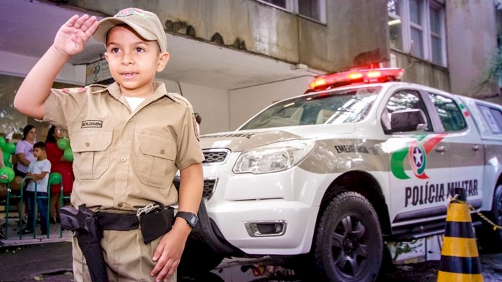 #PraCegoVer Na foto, um menino faz o sinal de sentido ao lado de uma viatura policial
