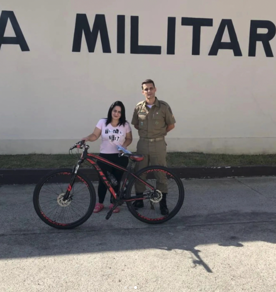 #PraCegoVer Na foto, uma mulher segura uma bicicleta ao lado de um policial militar