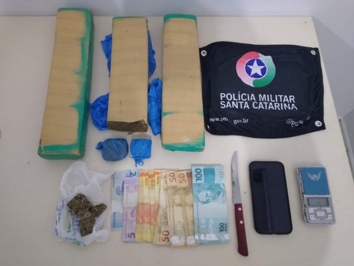 #PraCegoVer Na foto, uma bandeirola com o símbolo da Polícia Militar de Santa Catarina, tabletes de maconha, dinheiro e apetrechos utilizados para porcionar a droga.