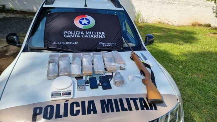 Drogas e arma em Criciúma #PraCegoVer Na foto, várias embalagens com drogas, uma espingarda de pressão, uma balança de precisão e duas facas estão em cima do capô de uma viatura da Polícia Militar
