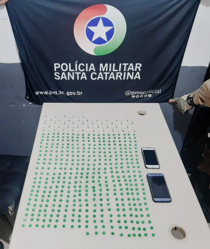 #PraCegoVer Na foto, Uma bandeira com o símbolo da Polícia Militar de Santa Catarina e vários comprimidos de ecstasy sobre uma mesa