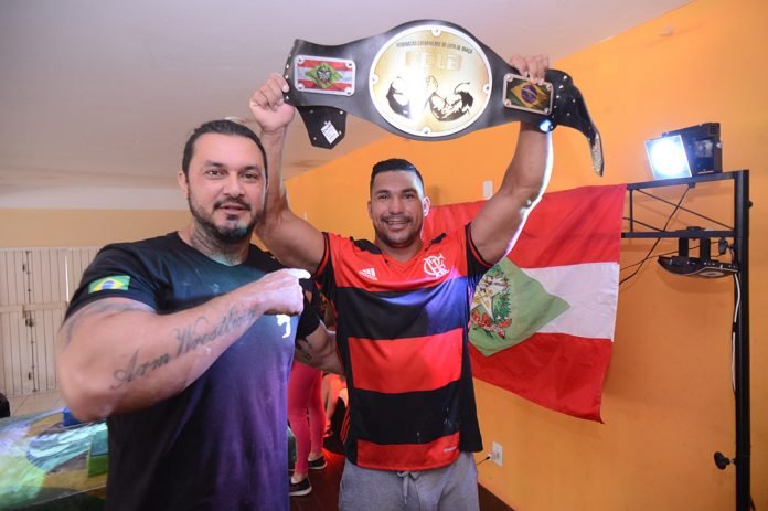 Nino queda de braço #PraCegoVer Na foto, um homem aponta para outro, que seguro um cinturão de ouro