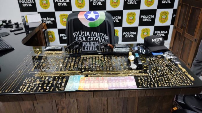 #PraCegoVer Na foto, uma mesa com vários produtos roubados. Ao fundo o símbolo da Polícia Civil e uma bandeira da Polícia Militar