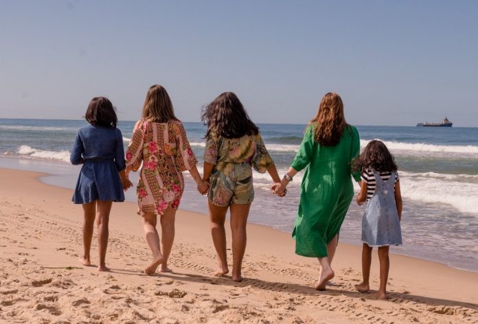 #ParaTodosVerem Na foto, cinco mulheres, duas delas adultas e as outras três menores, caminham em uma praia