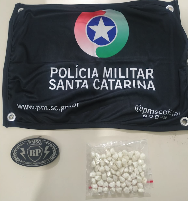 #ParaTodosVerem A foto mostra uma mesa e em cima dela há uma bandeirola com o símbolo da Polícia Militar, um pacote com pedras de crack e o brasão da Radiopatrulha da PM