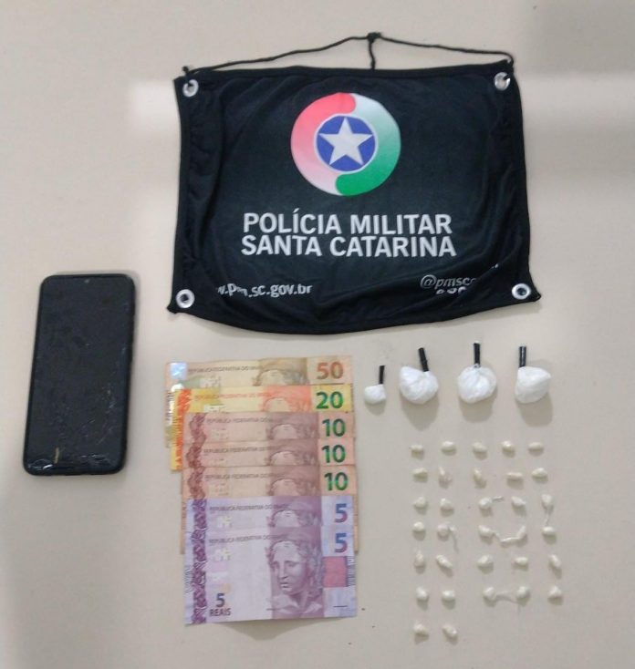 #ParaTodosVerem Na foto, em cima de uma mesa hão símbolo da Polícia Militar de Santa Catarina, um aparelho de telefone celular preto, notas de dinheiro e porções de cocaína e crack