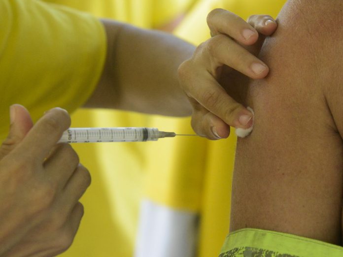 #PraCegoVer Na foto, uma pessoa com camisa amarela aplica uma injeção com vacina contra a gripe em outra pessoa
