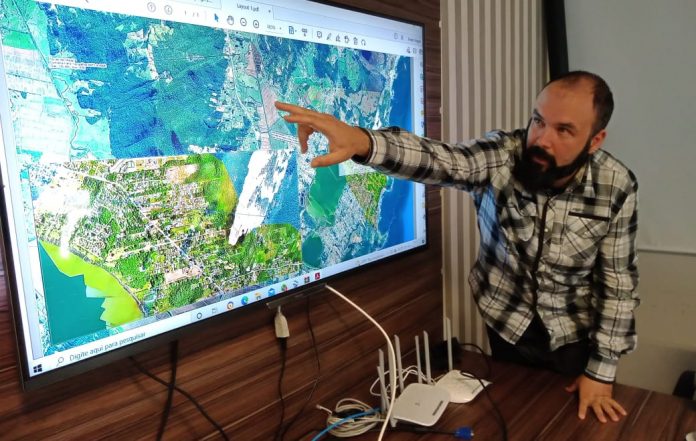 #PraCegoVer Na foto, um homem mostra o detalhe em um mapa cartográfico que é mostrado em um monitor
