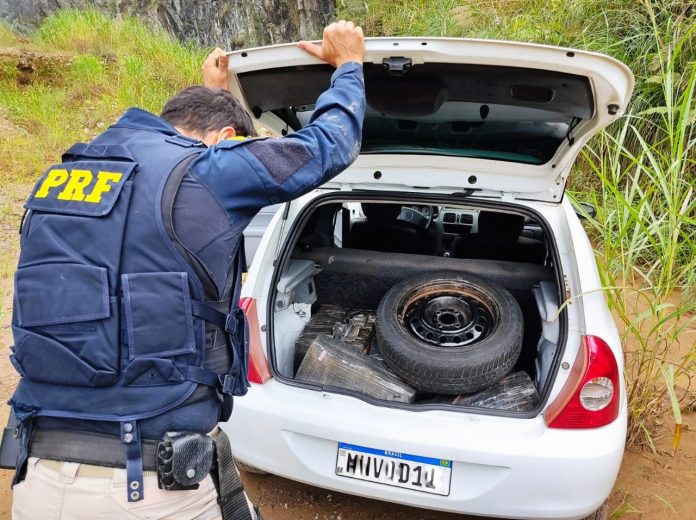 #PraCegoVer Na foto, uma agente da Polícia Rodoviária Federal inspeciona o porta-malas de um carro branco onde foi encontrado 215 quilos de maconha
