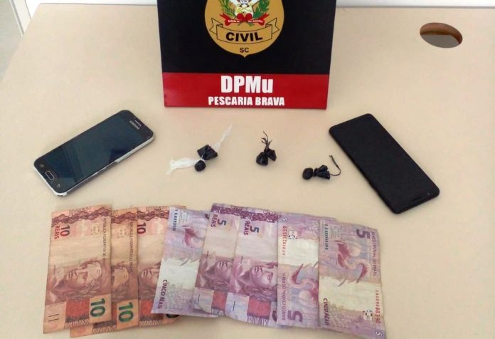 #ParaTodosVerem Na foto, uma placa com o símbolo da Polícia Civil, dois aparelhos de telefone celular, algumas cédulas de dinheiro e três porções de drogas