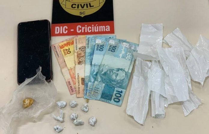 #PraCegoVer Na foto, uma mesa com o emblema da Polícia Civil, drogas, um aparelho de telefone celular e cédulas de dinheiro