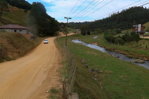 #ParaTodosVerem Na foto, uma estrada de chão batido. Ao lado há uma cerca de madeira e um riacho